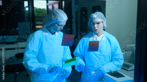 Two Female Scientists in Cleanroom Suits Examining Samples Under Blue UV Light in Modern Laboratory