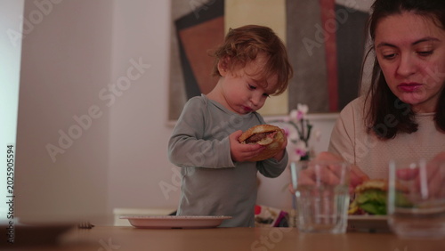 Fotografie Toddler standing at dining table holding burger with both hands, independent mea