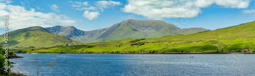 Green mountain slopes rise above Killary Fjord under bright skies in western Ireland. This image is a scenic landscape focused more on the fjord lush hillsides than the water.