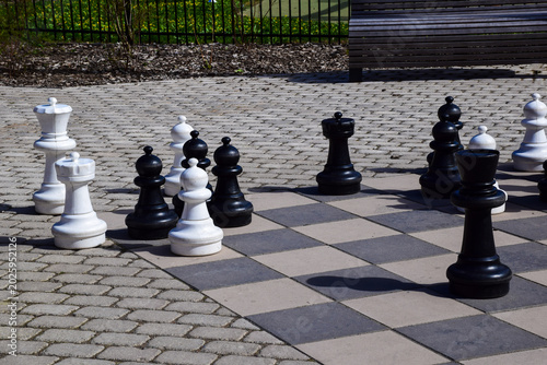  Large outdoor chess set with giant black and white pieces on a stone checkered board in a public park on a sunny day