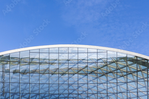 Curved glass facade displays repeating geometric patterns and reflective surfaces against a clear blue sky, highlighting modern architecture, transparency, symmetry and contemporary structural design