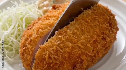 A close-up shot of a crispy golden Japanese pork cutlet being sliced with a knife, served with shredded cabbage on a white plate.