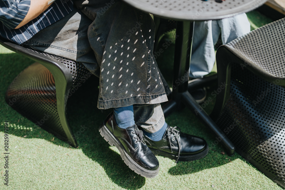 Fototapeta premium Two school friends sit with crossed legs under a round outdoor table, showing black shoes and denim. Casual students relax on patio chairs on artificial grass in a sunny outdoor setting.