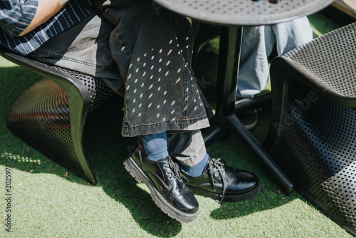 Two school friends sit with crossed legs under a round outdoor table, showing black shoes and denim. Casual students relax on patio chairs on artificial grass in a sunny outdoor setting.
