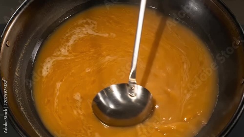 This close-up shot shows a metal ladle resting in a simmering orange savory soup inside a black cooking pot, capturing a home cooking scene.