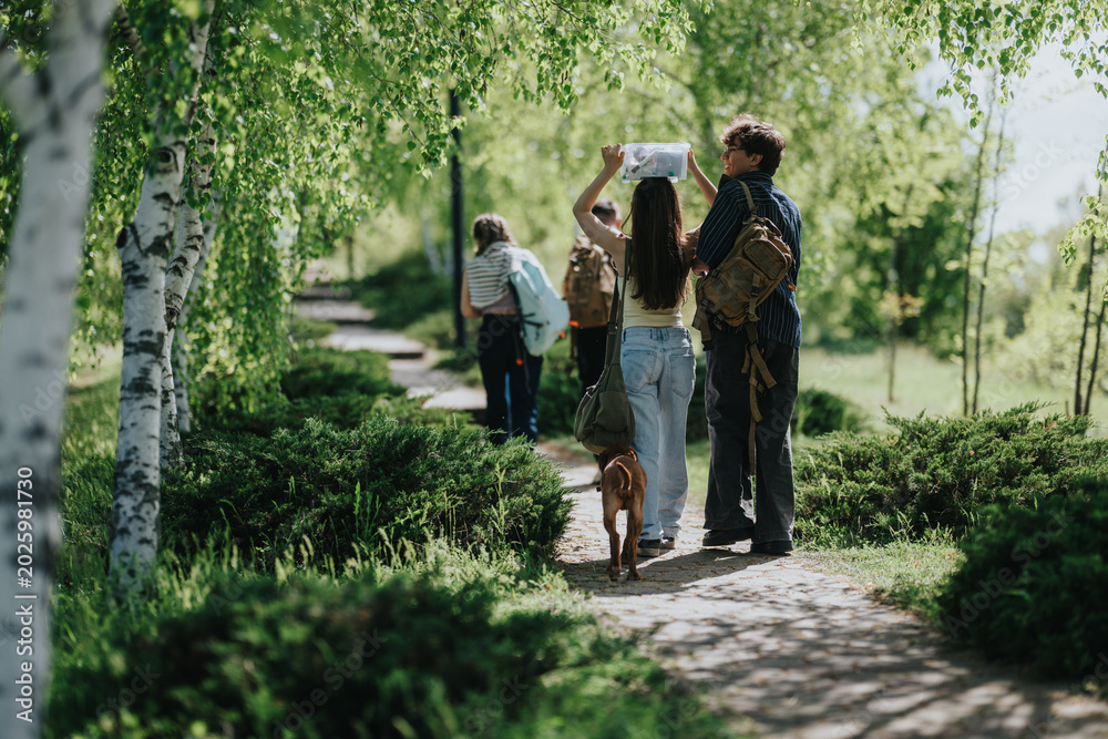Fototapeta premium A group of teenagers walk along a tree-lined park path with backpacks and a dog. They consult a map while strolling through green birch trees on a bright day.