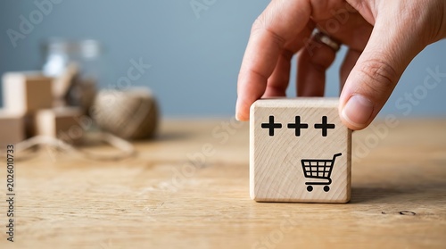 A hand placing a wooden block with a shopping cart and plus signs on a table with blocks and a jar in the background indoors.