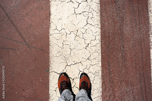Overhead view of feet in orange sneakers standing on cracked white street stripe between brown pavement. Urban texture and weathered road markings. For journey and decision concepts.
