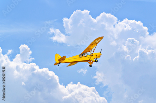 A yellow single-engine ultralight aircraft flying in a blue sky with white clouds.