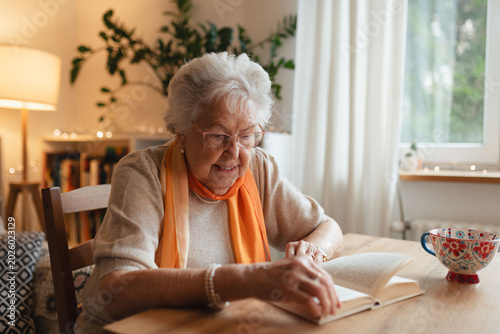 Photos Side view of elderly lady reading a book, sitting at the kitchen table and sipping tea
