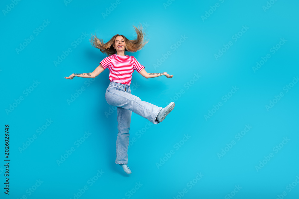 Fototapeta premium Joyful young woman in striped tshirt jeans jumps against blue background for lifestyle advertising and fashion promo