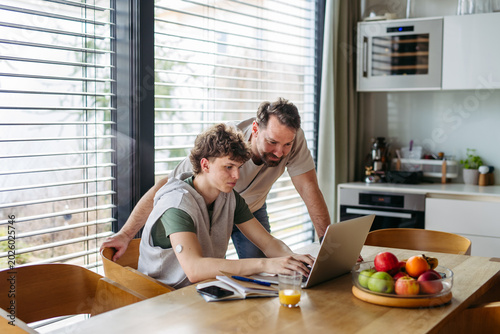 Foto Father helping teenage son with diabetes with homework on laptop in kitchen