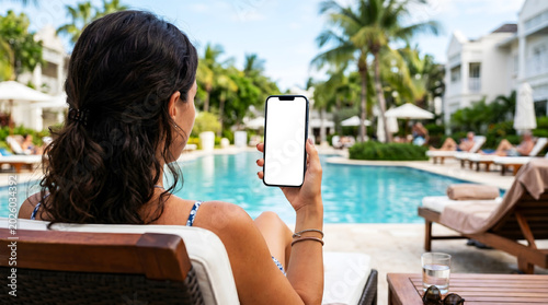 Hand holding mobile phone with blank white screen mockup, woman lounging by luxury tropical resort pool, summer holiday and digital travel lifestyle photography.
