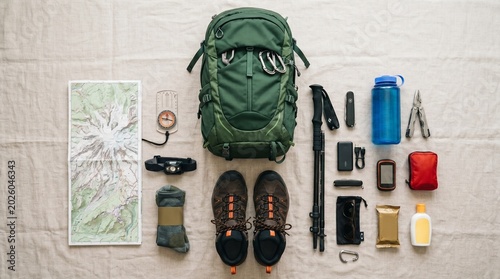 Flat lay of hiking gear and equipment for trekking, including a backpack, hiking boots, map, compass, water bottle, and survival tools on a light textured background
