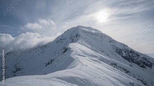 Group of hikers ascending a snow-covered mountain ridge under a bright sun and cloudy sky climbing