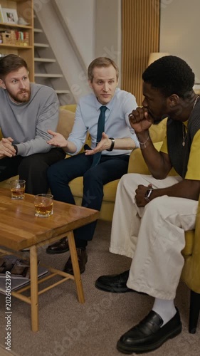 Vertical shot of biracial male friends gathering in living room, drinking bourbon and chatting, talking about family and life
