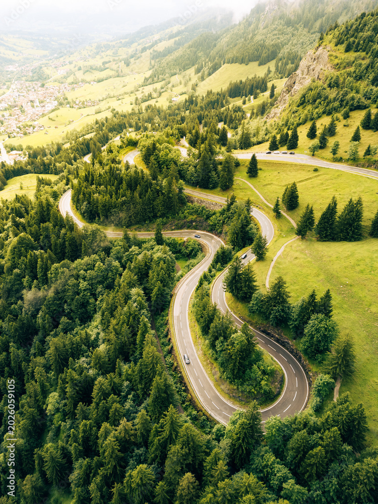 Obraz premium Travelers navigate a winding asphalt road through lush greenery in the Alps. The aerial view captures the curves of the serpentine path and surrounding hills during a sunny day.
