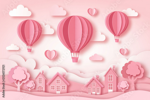 Paper art illustration featuring pink hot air balloons flying over cute suburban village landscape with clouds and hearts sky.
