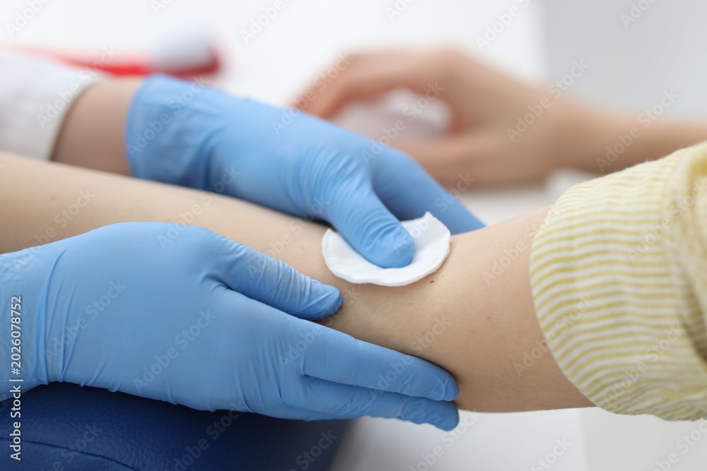 Fototapeta premium Nurse disinfecting patient's arm before drawing blood sample at white table in clinic, closeup