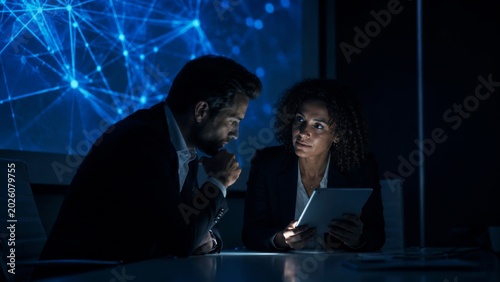 A dimly lit meeting scene showing two professionals engaged in discussion while examining a device, with a digital network backdrop illuminating the space.