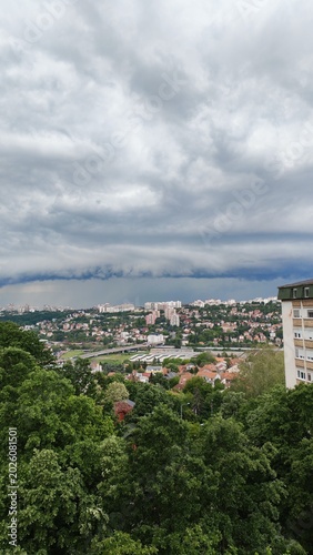 A dark storm cloud hanging over a city with buildings and forest in the background. Dramatic weather scene with contrast between urban area and nature.