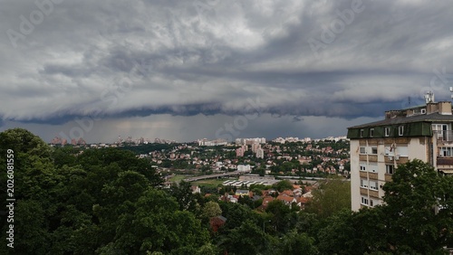 A dark storm cloud hanging over a city with buildings and forest in the background. Dramatic weather scene with contrast between urban area and nature.