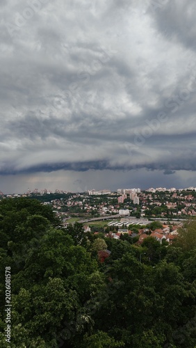 A dark storm cloud hanging over a city with buildings and forest in the background. Dramatic weather scene with contrast between urban area and nature.