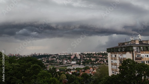 A dark storm cloud hanging over a city with buildings and forest in the background. Dramatic weather scene with contrast between urban area and nature.