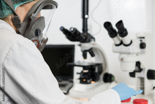 Obraz na plátně Over-the-shoulder view of a laboratory technician wearing a full protective mask and gloves while operating a modern microscope