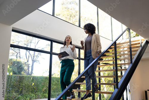Diverse female colleagues descending open-riser staircase, holding tablet, wearing office clothes