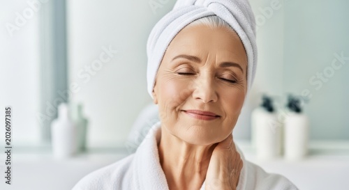 Smiling senior woman with towel on head and white robe touching neck in bathroom