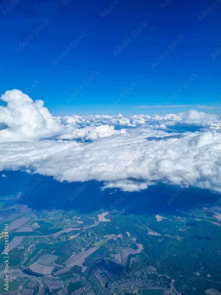 Obraz premium Aerial landscape view from airplane above dramatic white clouds and agricultural fields under bright blue sky. Scenic flight perspective showing countryside patterns and towering cloud formations from