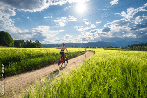 Vászonkép Woman riding mountain bike on dirt road through green wheat fields at sunset in Slovenia