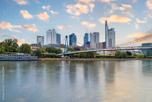 Skyline of Frankfurt's financial district, featuring prominent skyscrapers like the Commerzbank Tower, is reflected in the calm waters of the Main River, with the Holbeinsteg pedestrian bridge.