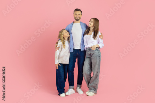 Canvas Print Happy caucasian family of three standing together on pink studio background, looking up father and smiling
