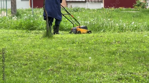 city utility worker or janitor in a blue coat and rubber boots cuts green grass and dandelions with a yellow lawn mower in the courtyard of an apartment building. Urban landscaping, spring cleaning, a