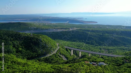 Panoramic aerial view of the lush Croatian coastline, featuring the iconic Krk Bridge spanning the Adriatic Sea, with winding roads and vibrant green hills. Highway A7