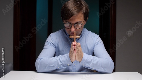 Canvas Print Young man with glasses bowing his head, holding a small wooden cross between pra