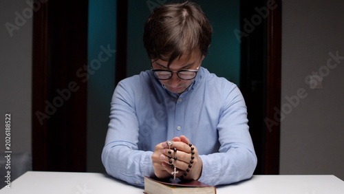 Bild auf Leinwand Man with glasses in blue shirt sits at a table, clasping rosary beads over an op