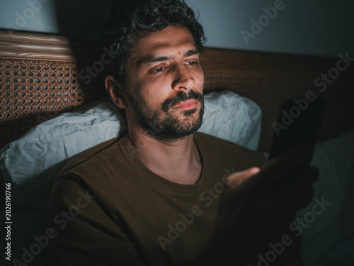 Man lying in bed at night, looking at his smartphone, suggesting themes of sleeplessness, technology use in the bedroom, or potential negative impact.
