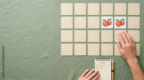 Woman playing memory game with matching apple cards on green tablecloth. Tabletop concentration board game with flat lay layout and score pad for recreational family home fun activity.