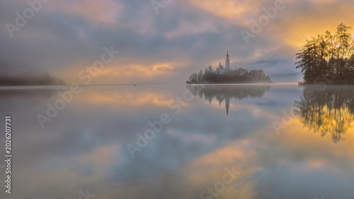 Tableau sur toile Reddish, thick fog sunrise over the Assumption of Mary church on Bled island, barely visible from Velika Zaka pier across Lake Bled