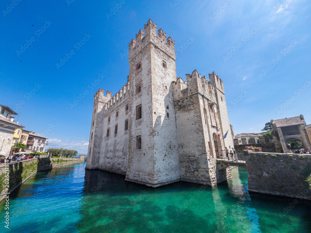 Obraz premium The historic Scaligero Castle in Sirmione rising directly from the crystal-clear, turquoise waters of Lake Garda on a bright, sunny day.
