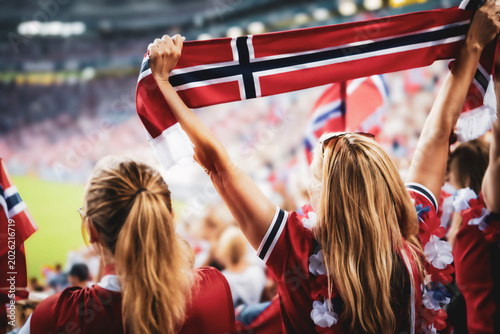 Back view of two female supporters holding the Norway national flag overhead during a soccer match in a crowded stadium atmosphere.