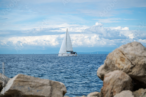 A monohull sailing yacht under party cloudy skies off the rocky coastline of Rovinj, Croatia.