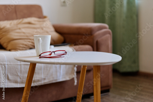 Obraz na plátne Cozy living room scene with a round white table, a mug and glass resting nearby, a soft sofa in the background, a warm natural light creating a calm home atmosphere soft textures in view