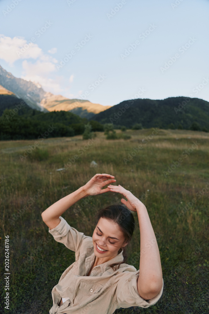 Fototapeta premium A happy woman in casual beige clothing enjoys nature with raised arms in a green meadow near forested hills under a clear sky. Lifestyle and outdoor concept.