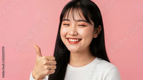 Joyful Expression: A young woman with long dark hair and a warm smile gives a thumbs-up, radiating positivity and happiness against a soft pink background.