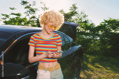 A woman with a retro hairstyle in a striped crop top stands beside a car  looking at her phone in a sunlit outdoor setting. Concept for casual style and modern connectivity.