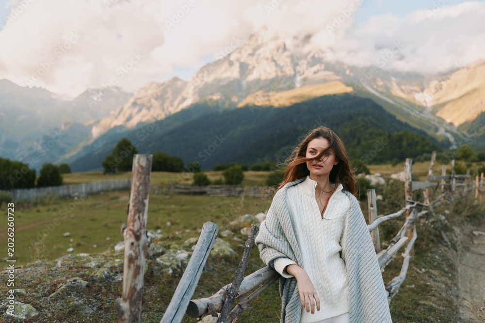 Fototapeta premium A young woman in a cozy knit poncho stands beside a wooden fence in a mountain valley, gazing ahead with a calm, reflective mood.
